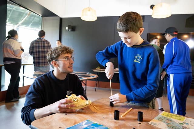 A child uses a wooden tool at a table while an adult, holding a skull model, observes in a workshop setting with others in the background.