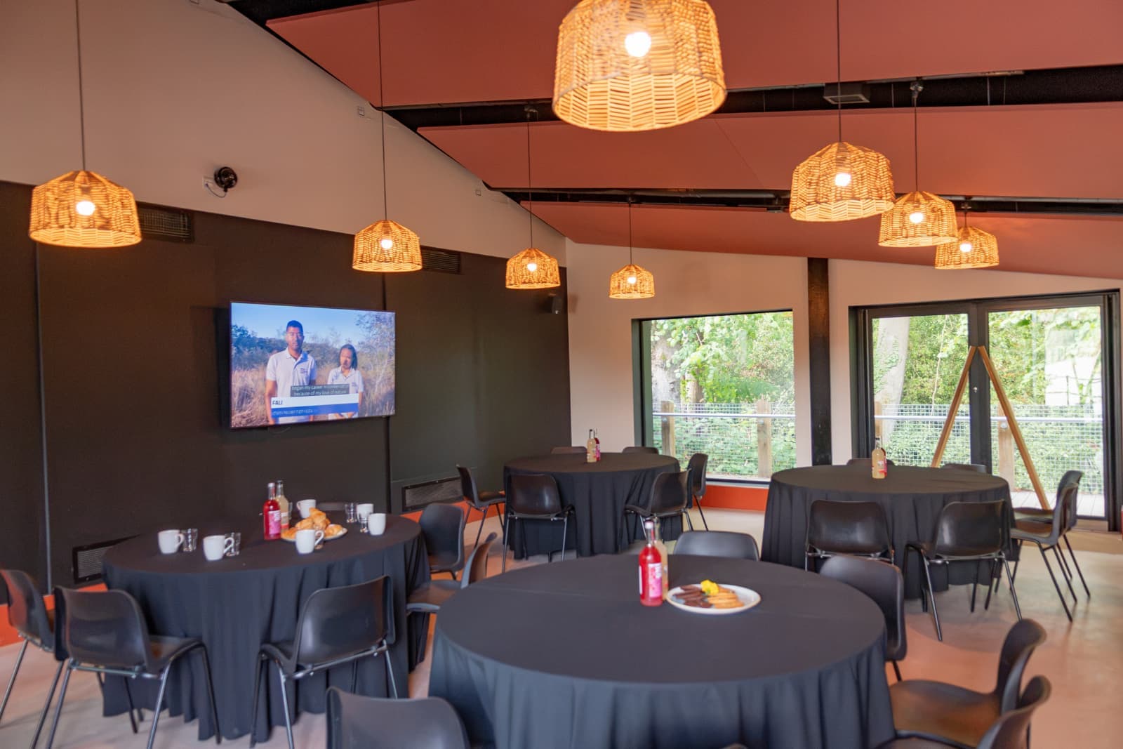 A cosy dining area with round tables covered in black tablecloths, wicker pendant lights, a TV on the wall, and a view of trees through large windows.