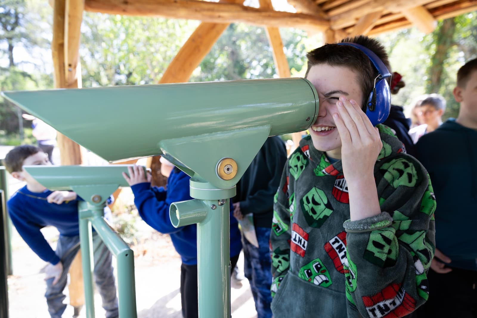 A boy wearing earmuffs smiles while looking through a large outdoor telescope, surrounded by other children.