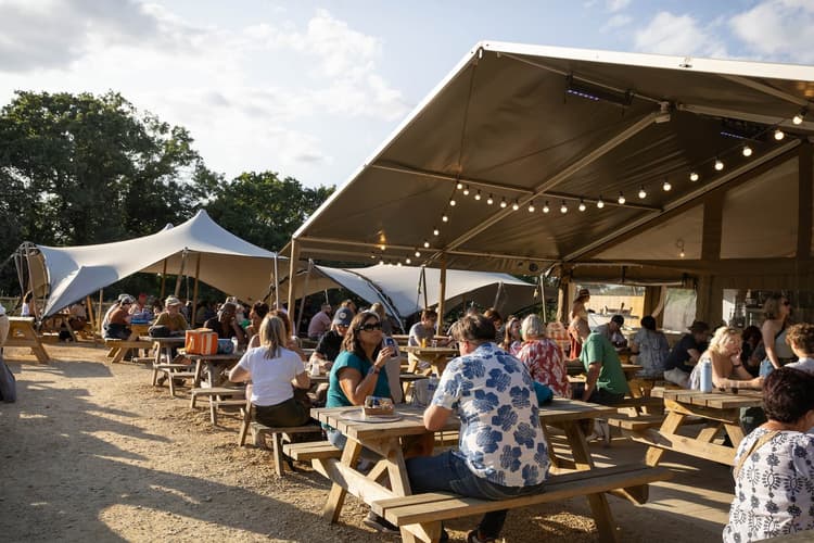 People sitting at picnic tables under shaded tents, enjoying a sunny day outdoors, with string lights hanging from the canopies.