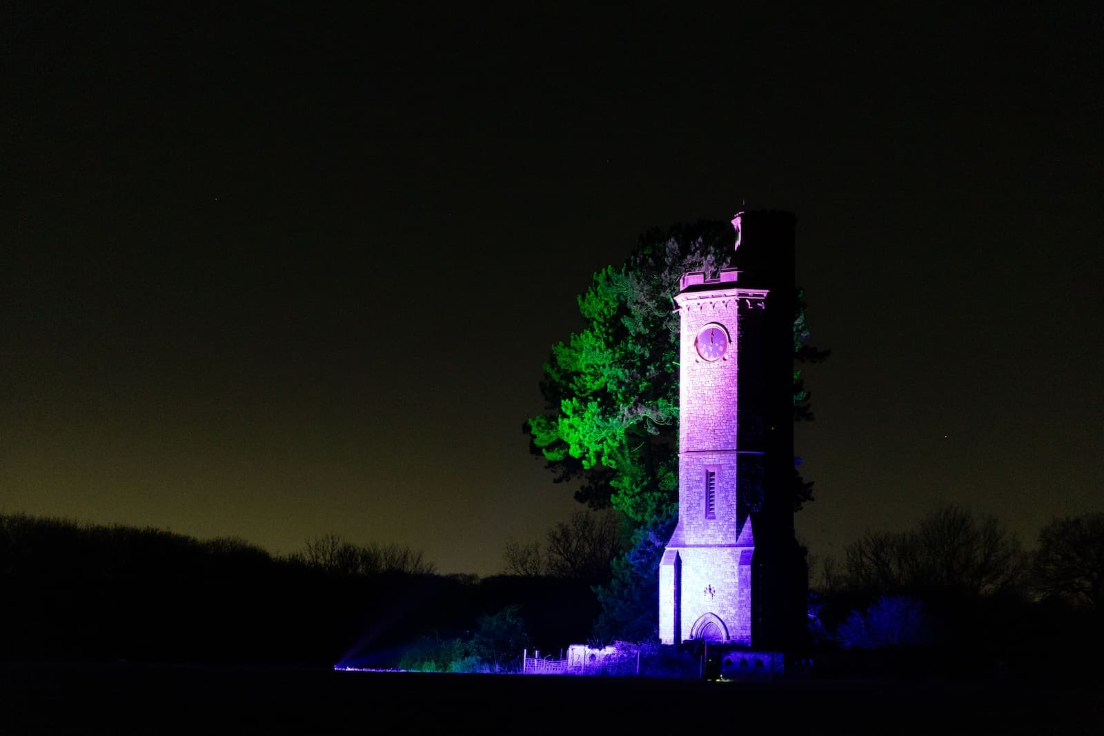Tall brick tower illuminated by colorful lights, surrounded by trees, set against a dark night sky.