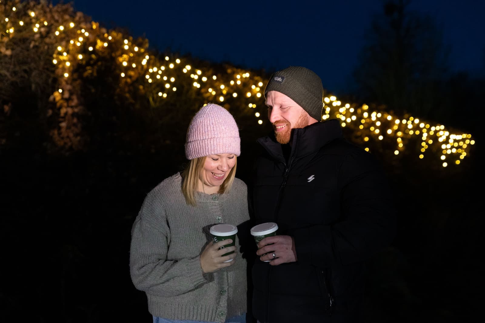 A couple in winter clothing smiles while holding hot drinks, with festive string lights glowing in the dark background.