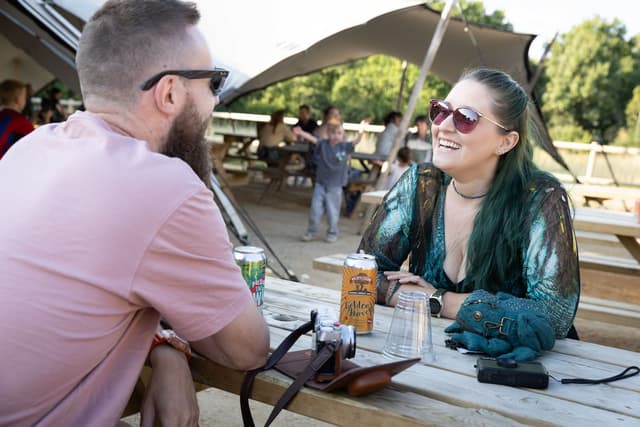 Two people sitting at a picnic table, smiling and talking, with canned drinks and cameras on the table, under a large tent in a park.