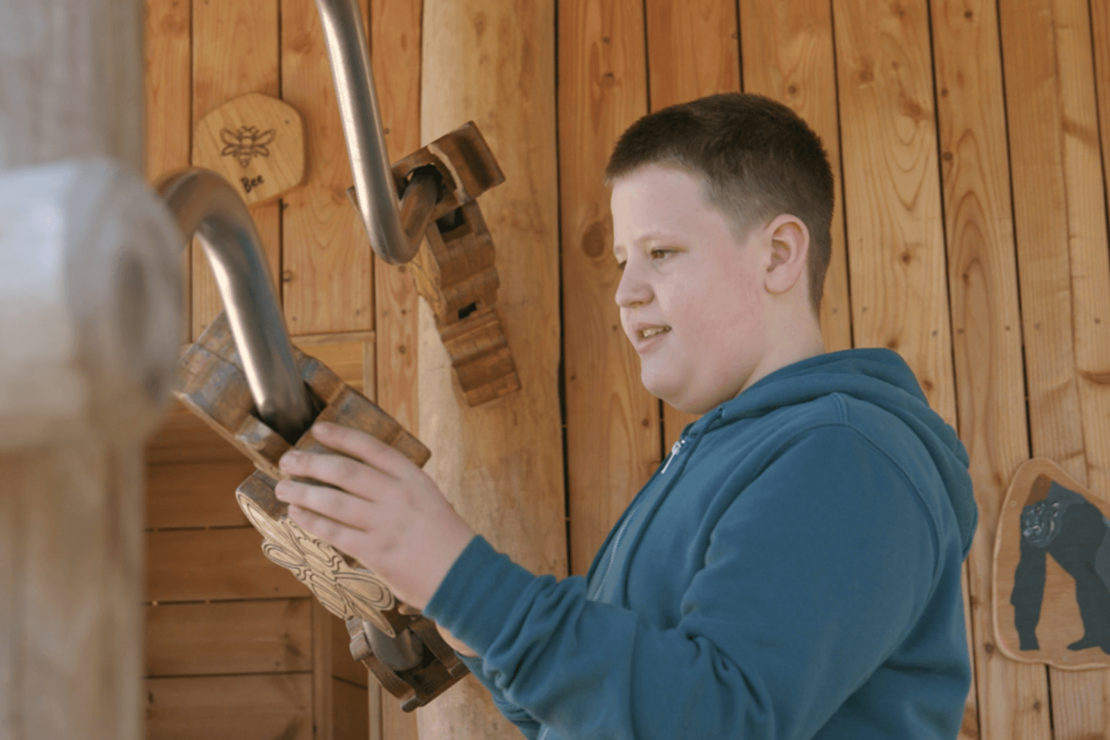 Boy in a blue hoodie interacts with wooden sculptures and metal tubes on a wooden structure, with a bee sign in the background.
