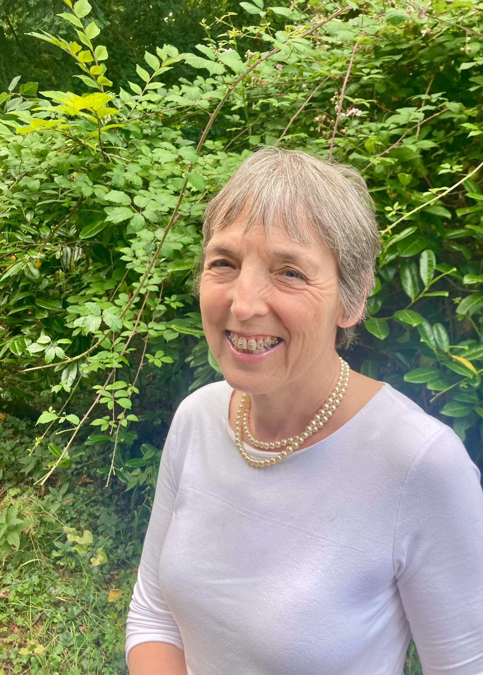 Woman smiling, wearing a white top and pearl necklace, stands in front of lush green foliage.