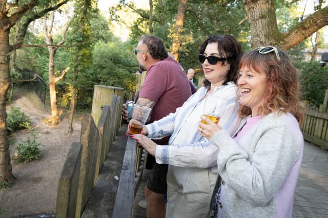 Three people enjoying drinks while standing on a wooden platform, overlooking a scenic, wooded area.