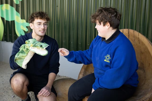 Two people interacting joyfully with a green crocodile puppet, sitting in a cozy, rustic setting with a wooden chair and green wall.