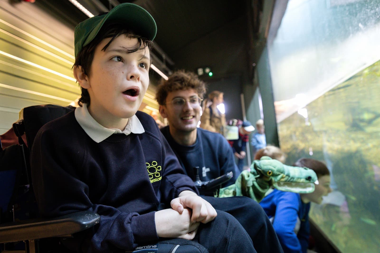 A child in a wheelchair wearing a cap looks in awe at an aquarium, accompanied by a smiling man and other children.