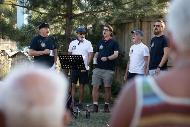 Five men singing outdoors with music stands, wearing casual clothes and caps, surrounded by trees and a wooden fence.