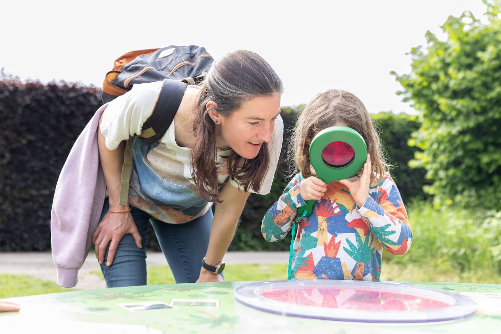 A woman and child explore outside; the child looks through a green magnifying glass while the woman leans in to watch.