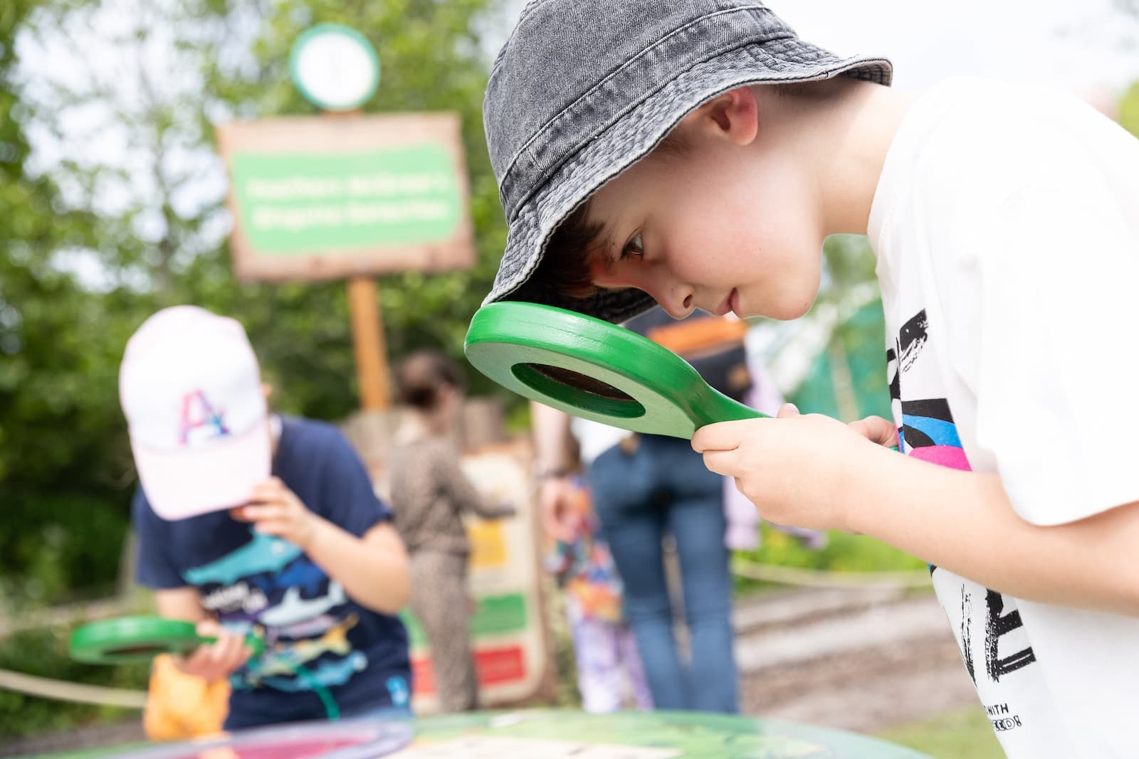 A child in a hat uses a green magnifying glass to examine something outdoors, with another child in the background doing the same.