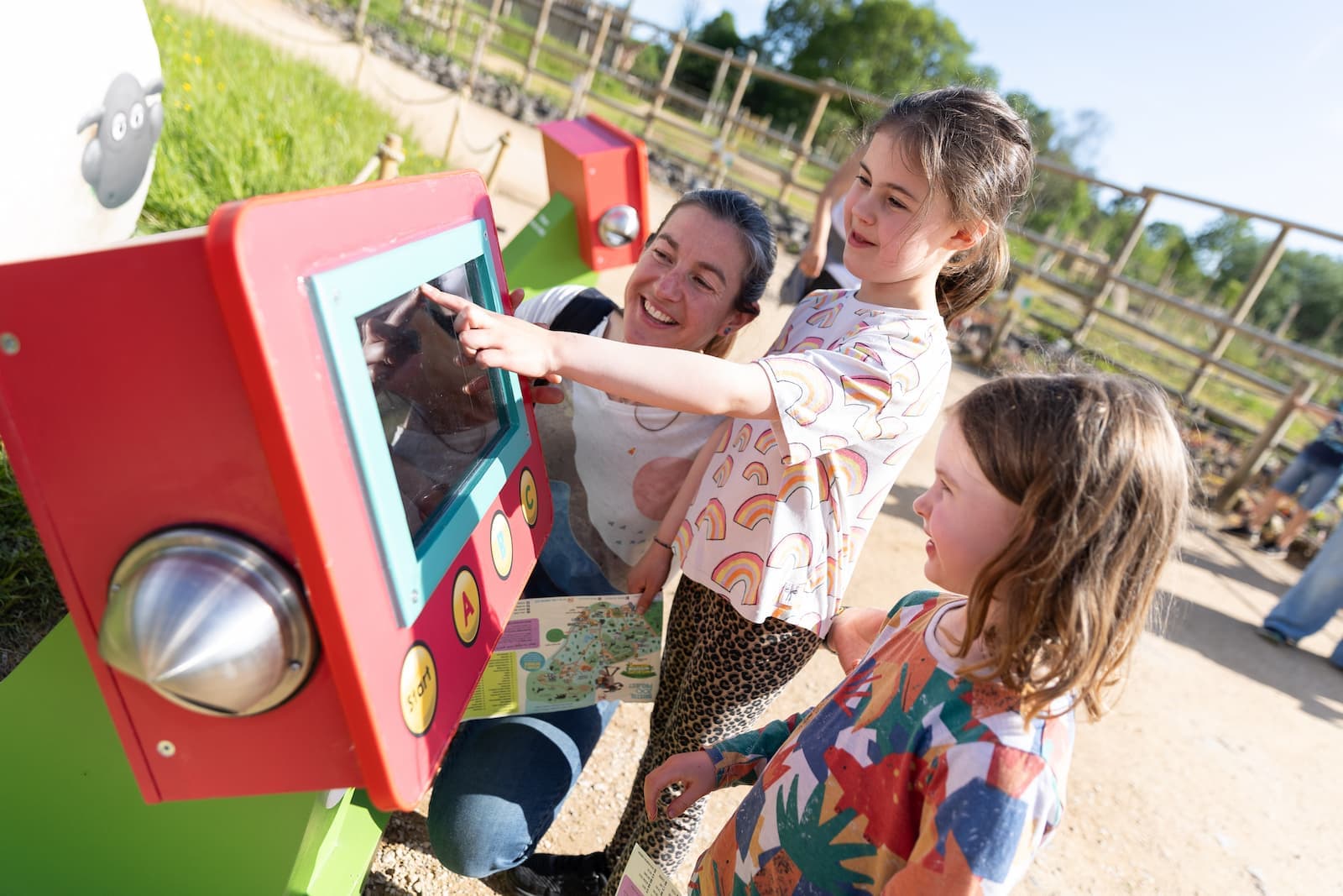 Two children and an adult interact with an educational touchscreen display outdoors, smiling and pointing at the screen.