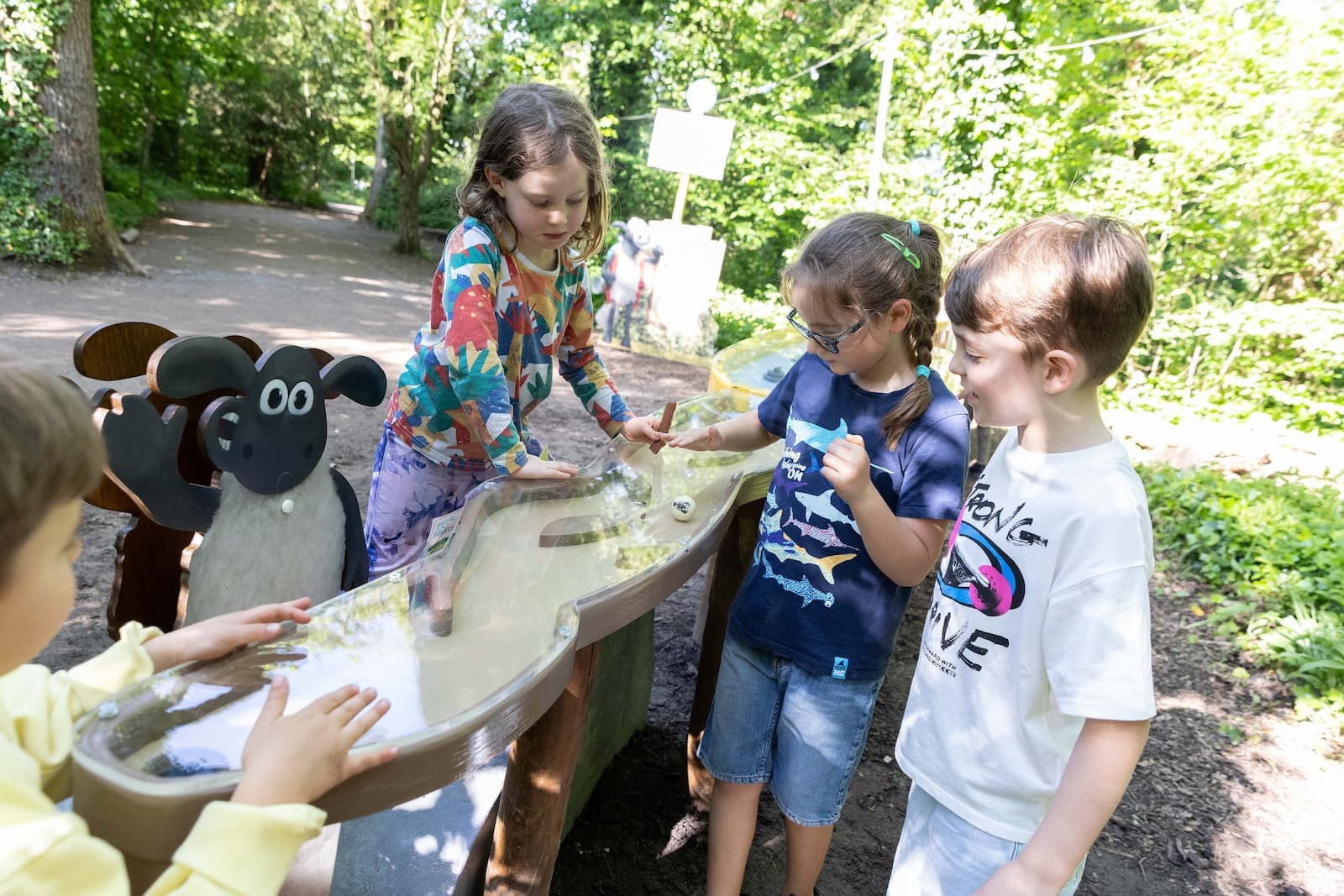 Children play with a water table in a park, interacting with a Shaun the Sheep figure, surrounded by lush greenery.