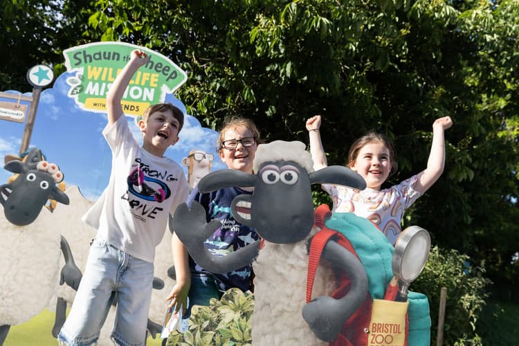 Three kids cheer excitedly beside a "Shaun the Sheep: Wildlife Mission" cutout display, outdoors on a sunny day.