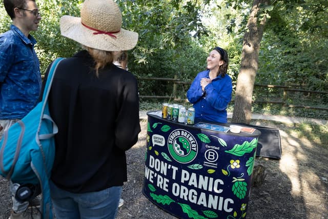 A woman in a blue shirt stands behind a table with organic beverage cans, talking to two people outdoors. The table banner reads "Don't Panic, We're Organic."