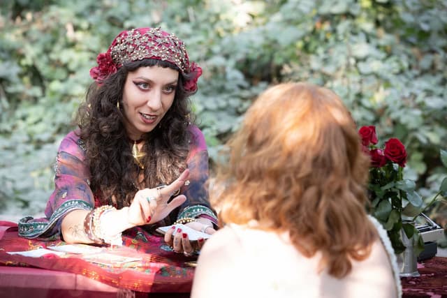 A fortune teller reads tarot cards for a woman in an outdoor setting, with roses on the side and sunlight filtering through the trees.