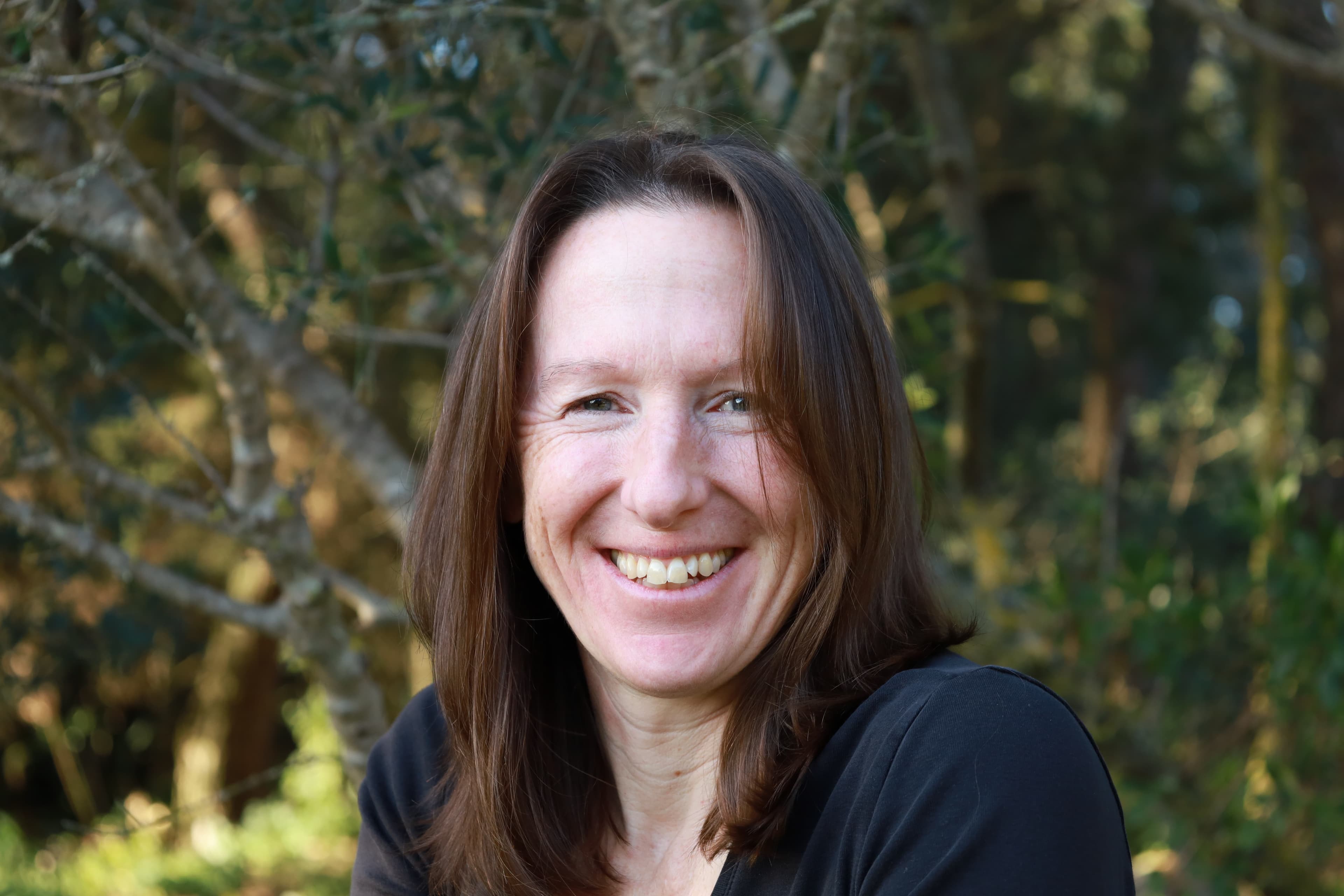 A woman with long brown hair smiles warmly, surrounded by a forest background.